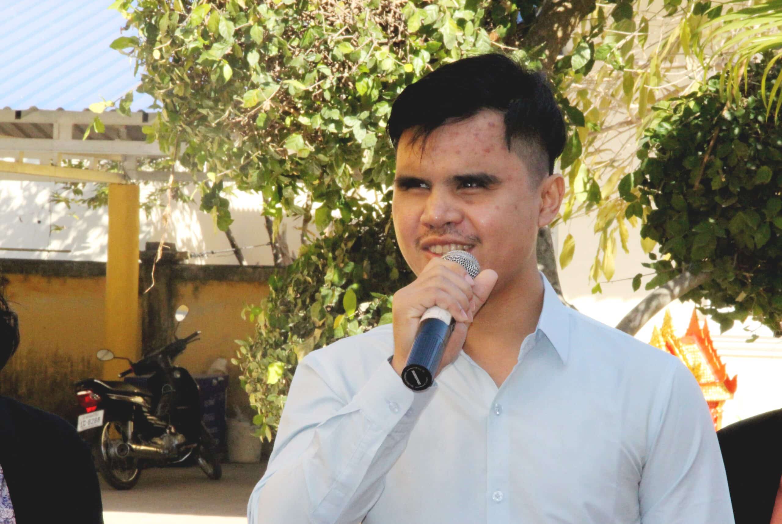 Vireak a young Cambodian man holds a microphone giving a speech in a garden.