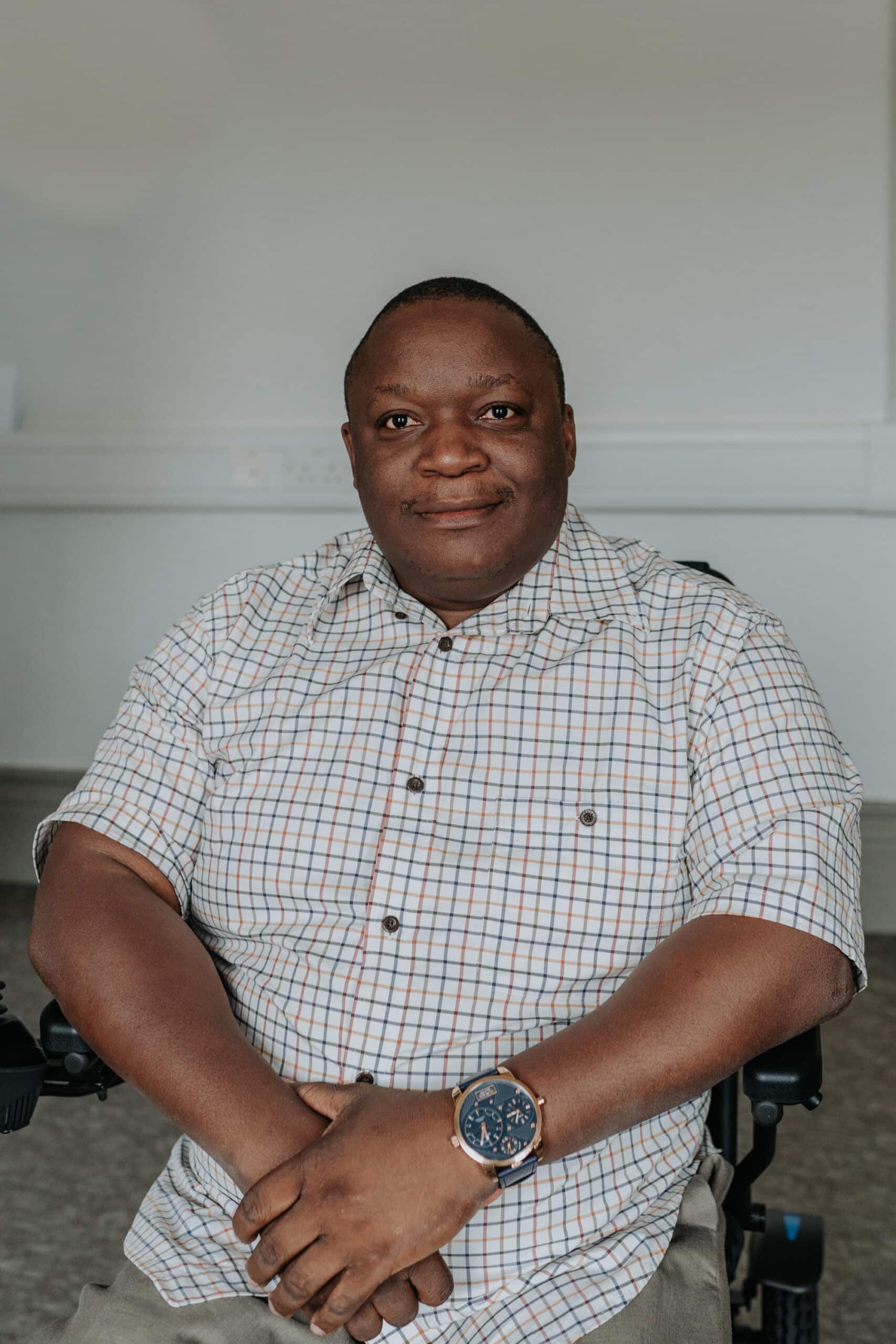 Fredrick sitting in his wheelchair in an office room smiling to camera wearing a light checkered shirt.