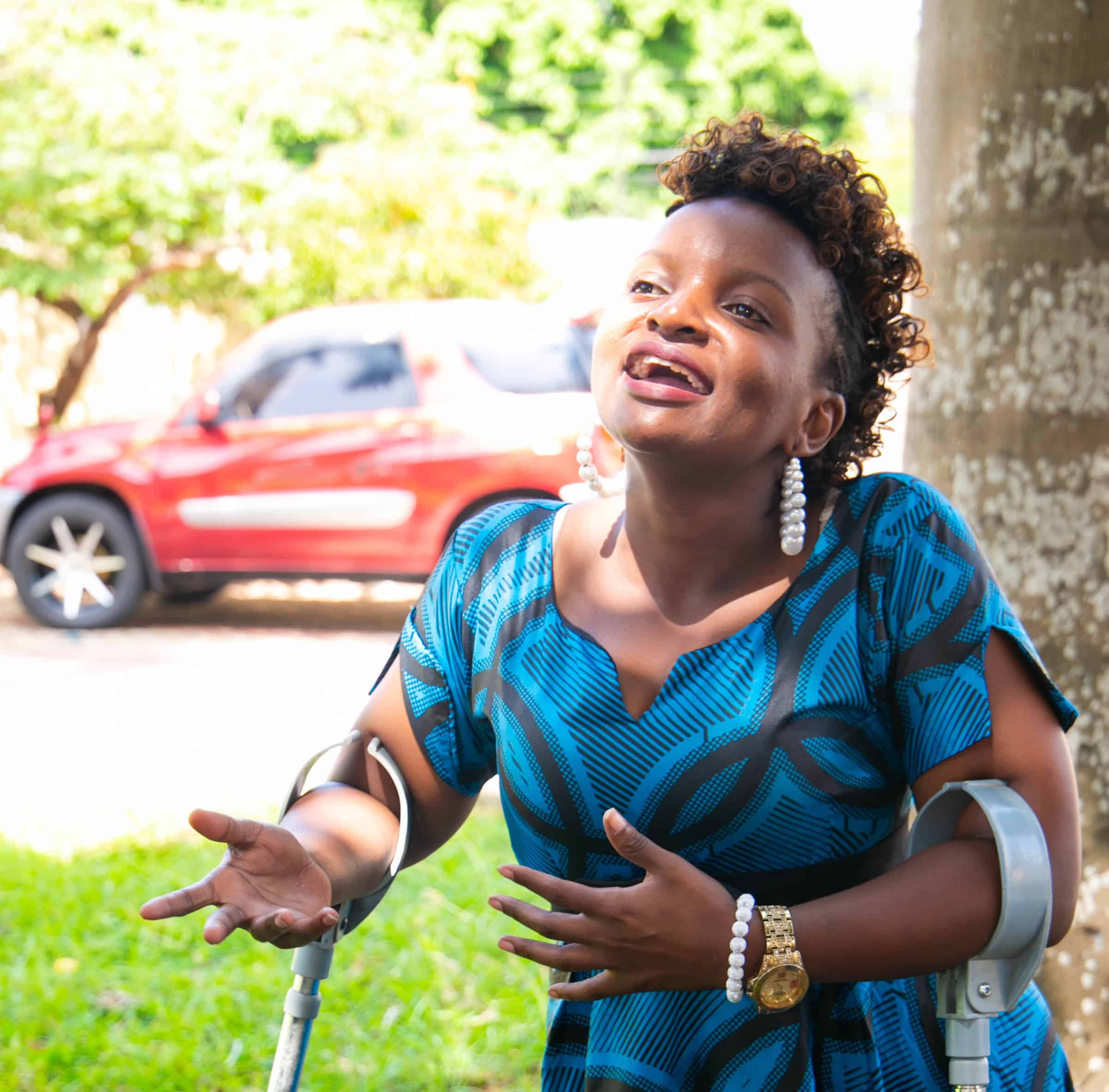 Dorice is speaking and gesticulating with her hands. She wears a blue dress and stands using crutches.