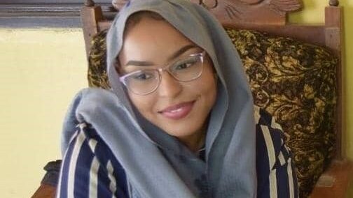 Elkhansa wears a blue striped top and a grey headscarf and glasses. She is smiling to the side of the camera.